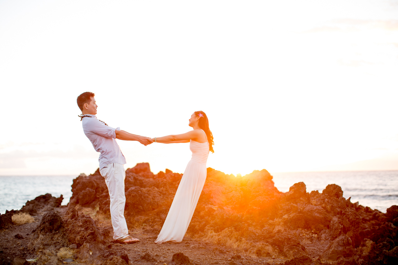 A couple leans back holding hands as a sunset glows over the rocks and ocean.