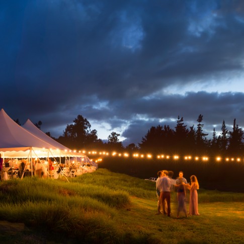 A photo of a wedding reception in Kula Maui with a white tent and the glow of lights while the sky turns dark.