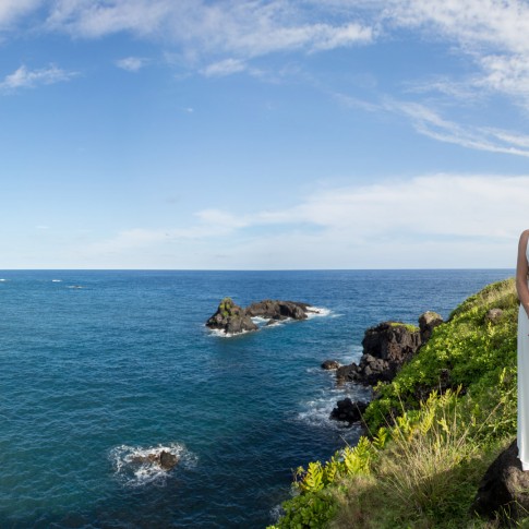 A bride stands alone on the edge of a cliff set against the blue ocean at a destination wedding in Hana Maui.