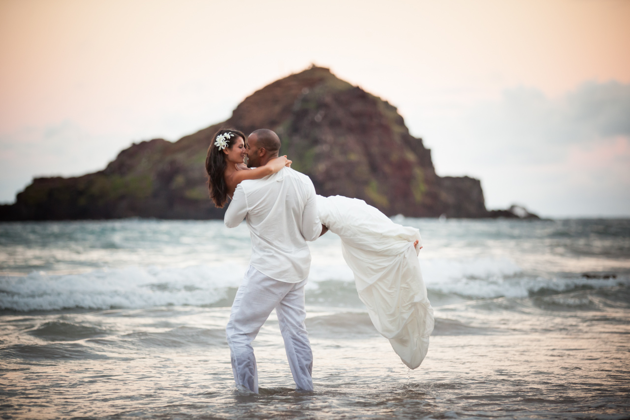 A groom holds his bride at Koki beach in Hana Maui with Alau Island in the background.
