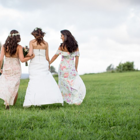 A bride and her bridesmaids walk away from the camera across a field as their dresses fly in the wind in Hana maui.