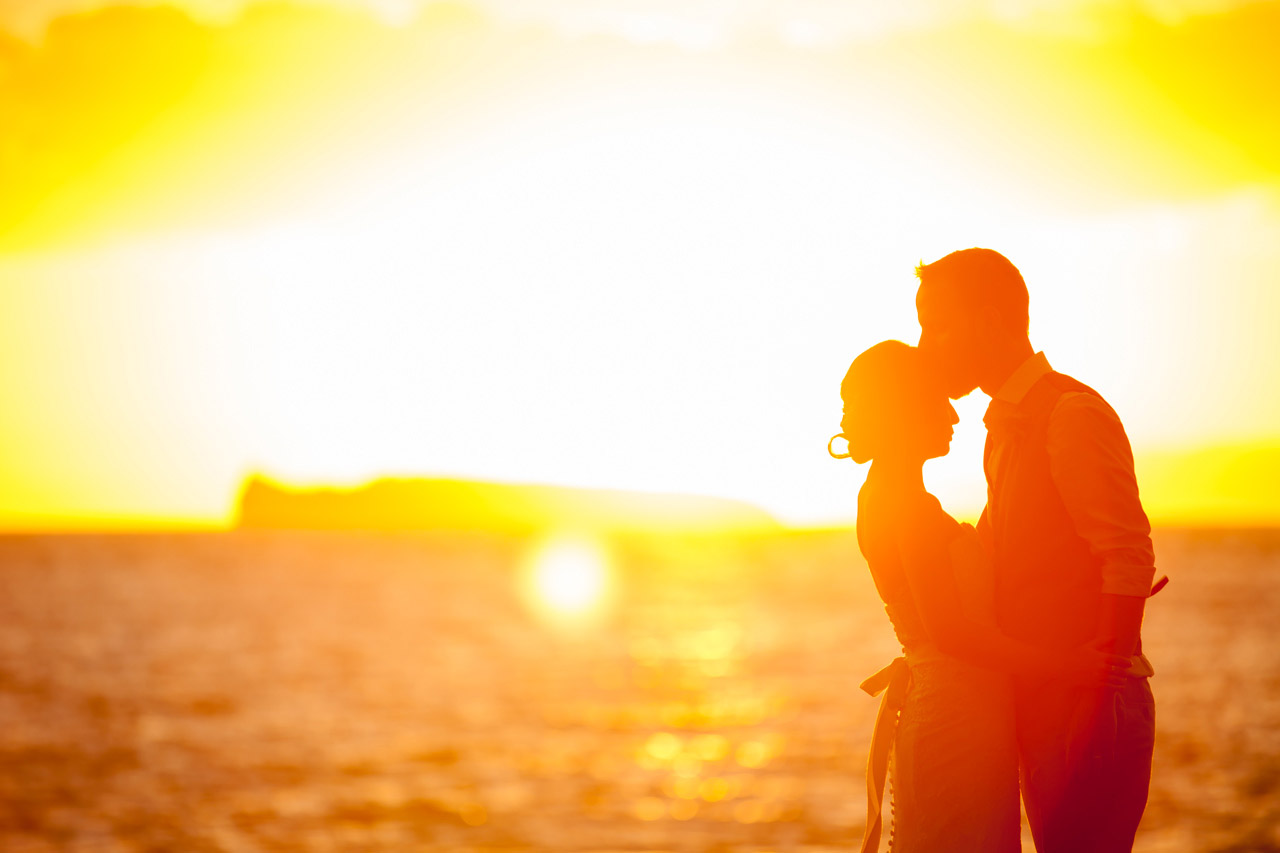 A groom kisses his brides forehead as a dramatic sunset flares in the background at a destination wedding in Hawaii.