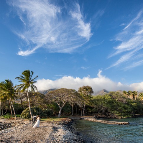 A couple walk along the ocean as whispy clouds and tropical mountains loom overhead in Olowalu, Maui.