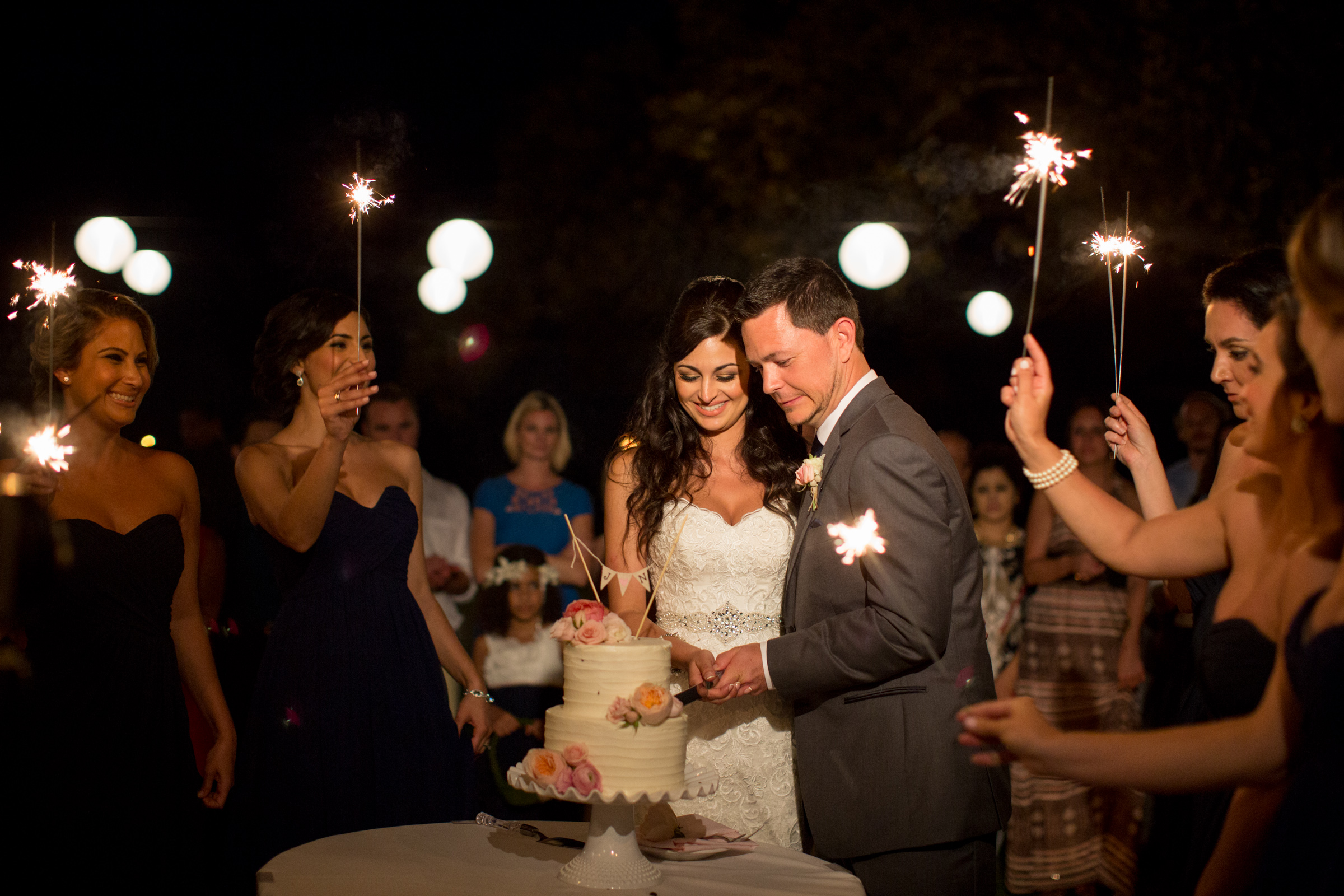 A couple cut their cake at their wedding freception while guests look on with sparklers.