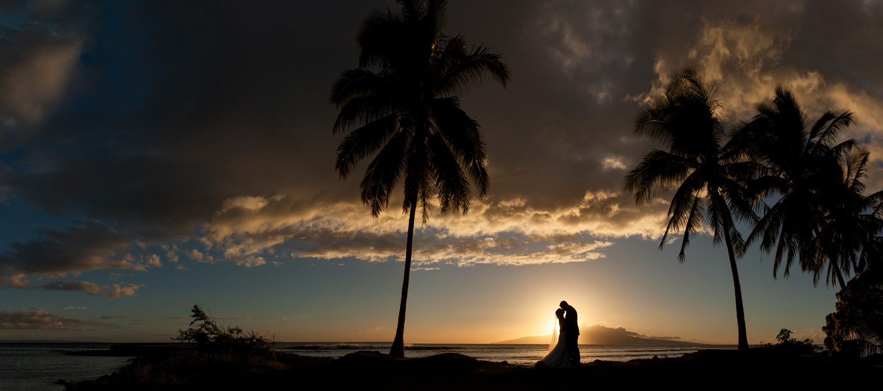 A couple silhouetted against a palm tree liined sunset in Maui.