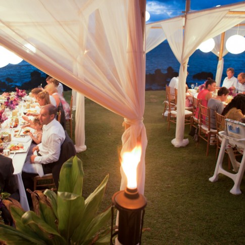 A photo of 2 tables of weddings guests enjoying the sunset dinner with tiki torches.
