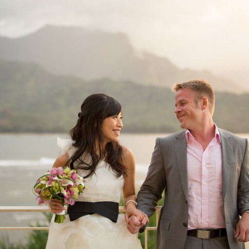 a picture of a bride and groom leaning against a railing set against the hanalei bay on kauai