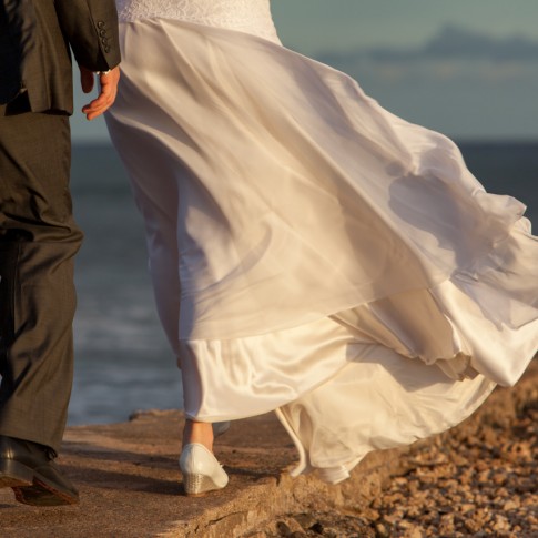 a brides dress flows in the wind as a wedding coouple walk along a seawall