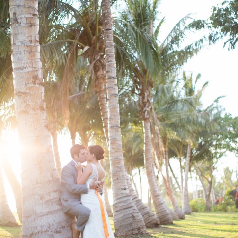 Photo of a wedding couple kissing while leaning against a row of palm trees