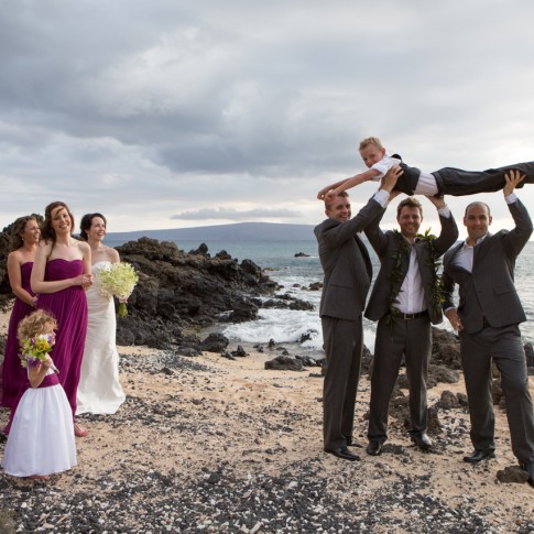 Groomsmen hold a ring bearer above their heads while the bride and bridesmaids watch .