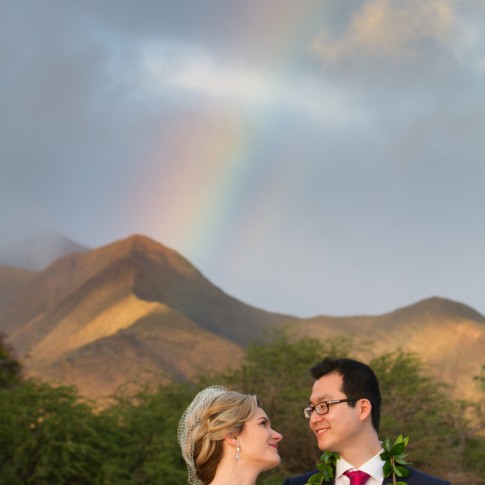 A wedding couple gaze into each others eyes as a rainbow descends above them.