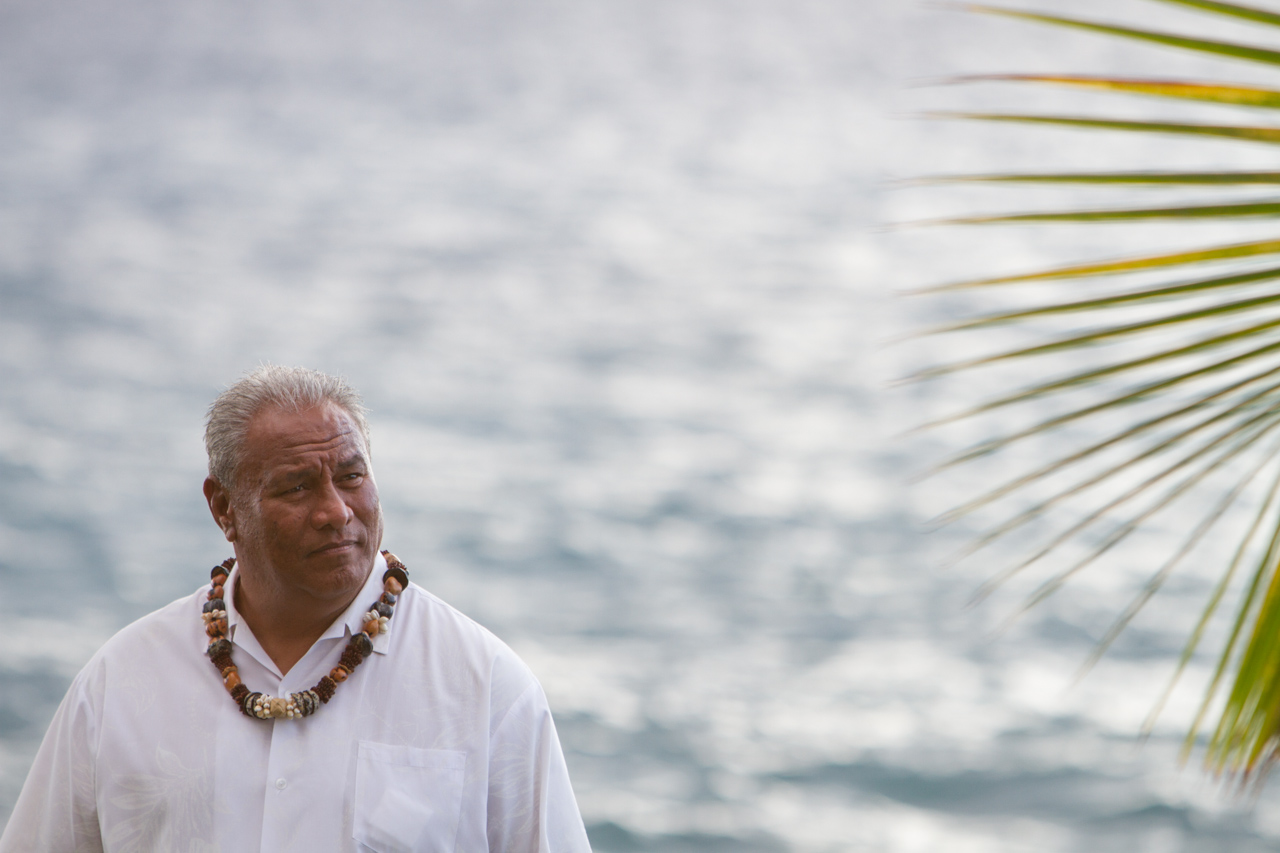 Photograph of the Minister wearing a kukui lei with palm fronds and the ocean in the background.
