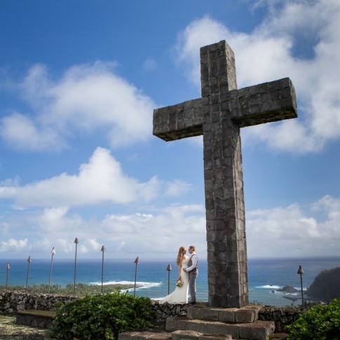 Bride and groom embrace as they stand at the base of Fagan's Cross overlooking Hana Town and the turquoise ocean
